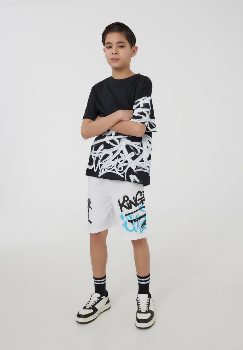 T-shirt nera con design grafico bianco su una manica, abbinata a pantaloni corti bianchi con testo graffiti nero e blu. Sneakers in bianco e nero.