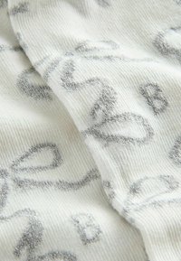 Tejido de punto blanco con lazo plateado brillante y patrones de la letra B, mostrando textura y pliegues en detalle de cerca.