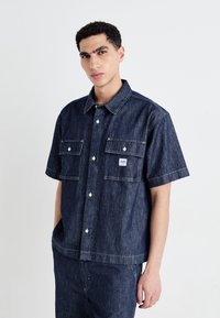 Dark denim short-sleeve shirt with two front pockets, button closure, contrasting stitching, and a small logo patch on the left chest.