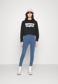 Črna kapuca z logotipom "Levi's", modri oprijeti džinsi, beli visoki športni copati in bež vedro klobuk, postavljeni na enobarvni beli ozadju.