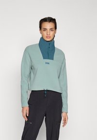Cuerpo recortado de forro polar azul claro con cuello alto, media cremallera y logo. Cordón ajustable en el dobladillo. Combinado con pantalones cargo oscuros.