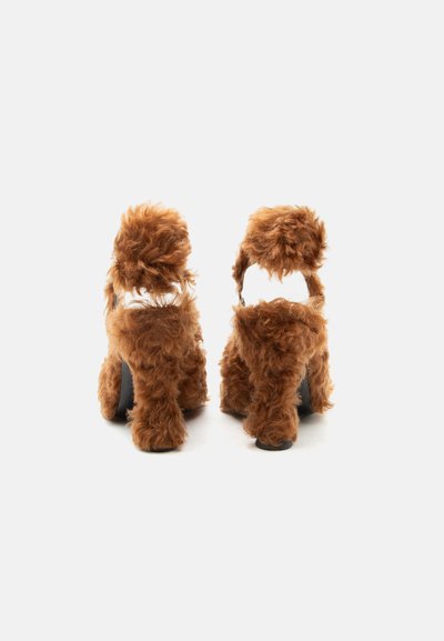 Καφέ αφράτα πέδιλα με ψηλό τακούνι και λουριά στον αστράγαλο, καλυμμένα με μαλακή συνθετική γούνα, εμφανιζόμενα από πίσω σε λευκό φόντο.