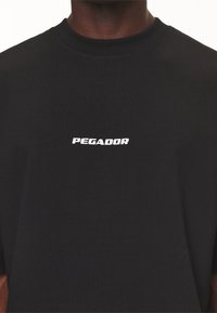 Svart bomullströja med en vit "PEGADOR"-logotyp i fet stil, rund halsringning, korta ärmar och en mjuk textur.