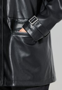 Giacca in pelle nera con una texture liscia, tasche laterali e fibbia regolabile sul polsino. Design semplice con linee pulite.