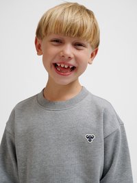 Uśmiechnięty młody chłopiec o blond włosach i przerwach między zębami z przodu, ubrany w szarą bluzę z małym logo pszczoły na piersi.