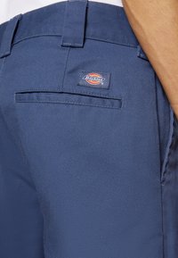 Gros plan sur la poche d’un pantalon Dickies bleu marine avec un écusson logo au-dessus de la poche arrière passepoilée sur la hanche d’une personne.