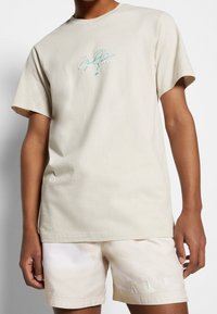 Beige puuvillainen t-paita lyhyillä hihoilla, jossa on turkoosia tekstiä ja allekirjoitusgrafiikkaa edessä. Sujuva pinta, rento istuvuus.