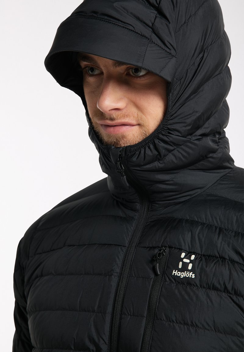 Chaqueta negra acolchada con un cuello alto y capucha adjunta. Presenta una cremallera y un logo en el pecho. Tejido suave, aislante y con un patrón acolchado.