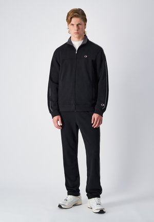 Veste de survêtement zippée noire avec des rayures blanches et un logo sur la poitrine, associée à un pantalon assorti et des baskets blanches. Tissu lisse et chaud.