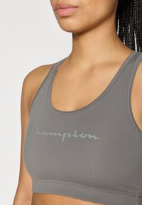 Γκρι αθλητικό σουτιέν με στρογγυλεμένο ντεκολτέ, λογότυπο σε ανοιχτότερο γκρι, κατασκευασμένο από λείο υλικό, με εφαρμογή που αγκαλιάζει το σώμα και απλές ραφές.