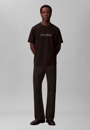 Mladý muž stojaci so založenými rukami za chrbtom, oblečený v čiernom tričku Calvin Klein, čiernych nohaviciach a čiernych topánkach na sivom pozadí.