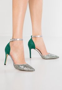 Högklackade skor med en glittrande silvertå och gröna mockasidor, med vristremmar, mot en vit bakgrund.