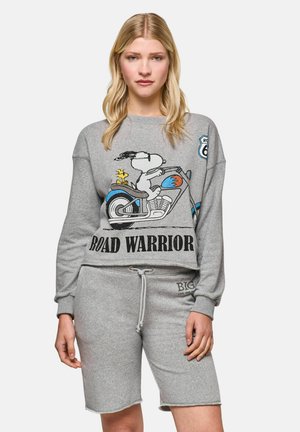 Woman with blonde hair wears a grey sweatshirt featuring Snoopy on a motorcycle and the words "ROAD WARRIOR," with matching grey shorts.