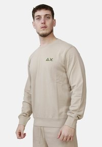 Jonge man in een beige sweatshirt en bijpassende broek, staand tegen een effen lichte achtergrond, iets naar links gekeerd.