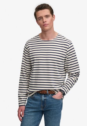 Mann mit braunen Haaren, der ein weißes und marineblau gestreiftes Langarmshirt, blaue Jeans, einen braunen Gürtel und eine schwarze Armbanduhr trägt, mit der Hand in der Hosentasche.