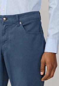 Ciemnoniebieskie chinosy o gładkiej fakturze, z przednią kieszenią, srebrnym guzikiem i kontrastowym przeszyciem. Połączone z jasną koszulą w krate.