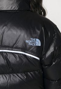 Czarna kurtka puchowa o błyszczącej fakturze, z niebieskim logo "The North Face" i białym poziomym paskiem na klatce piersiowej.