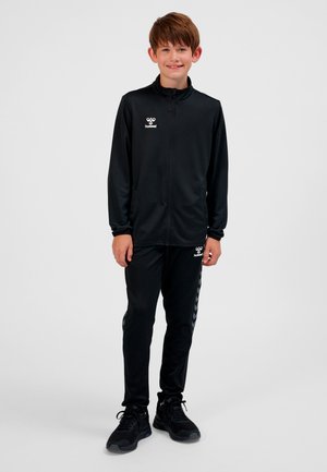 Schwarzer sportlicher Trainingsanzug mit Reißverschlussjacke und Hose, versehen mit einem Logo auf der Brust und seitlichen Chevron-Muster-Akzenten. Getragen mit schwarzen Turnschuhen.