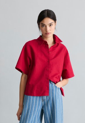 Junge Frau mit dunklen Haaren trägt ein knallrotes kurzärmeliges Hemd mit Knöpfen und hoch taillierte blaue gestreifte Hose, posiert mit der Hand in der Tasche.