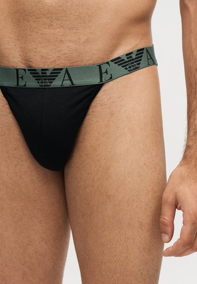 Gros plan d'un homme portant un sous-vêtement noir avec une ceinture verte comportant des lettres noires et des logos d'aigle.