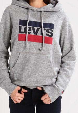Personne portant un sweat à capuche gris Levi’s avec un logo bleu marine et rouge, les mains tenant l’ourlet du sweat, et un pantalon noir.