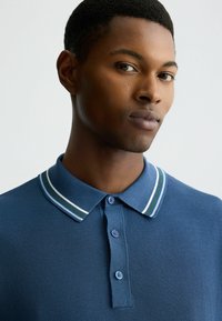 Blaues Polohemd mit strukturiertem Stoff, drei Knöpfen und einem Kragen mit weißen und grünen Streifenelementen. Nahaufnahme des Ausschnitts.