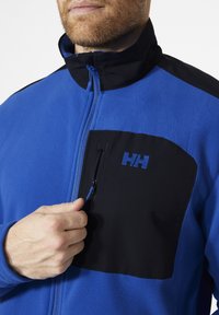 Fleecejacke mit einem blauen Oberteil und einer schwarzen Reißverschlusstasche auf der Brust. Blauer Reißverschluss-Zip und Logo auf der Tasche in passendem Blau.