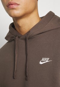 Brun Nike-hoodie tillverkad av mjukt material, med en framficka i kängurustil, vitt broderat logotyp och justerbar dragsko på huvan.