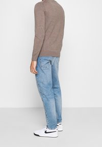 Mann in hellblauen Jeans, weißen Nike Sneakers mit schwarzem Swoosh und einem braunen Langarmpullover, der vor einem weißen Hintergrund steht.
