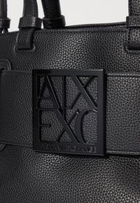 Musta tekstuuripinta nahkakotelo, jossa on suuri musta logo-levy, jossa on kohokuvioitu "ARMANI EXCHANGE" ja geometriset aukot.