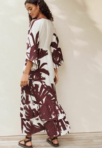 Langes, fließendes Kleid in Weiß mit dunklem roten Palmblättermuster. Verfügt über weite Ärmel, gestufte Rüschen am Saum und eine geraffte Taille.