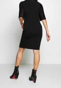 Czarna dopasowana sukienka z krótkimi plisowanymi rękawami, do kolan, zestawiona z czarnymi botkami za kostkę z kontrastową czerwoną podeszwą i masywnym obcasem.