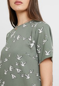 Woman wearing a green short-sleeve top with a white bird print and a delicate necklace, showing neckline and upper torso.