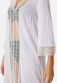 Cover-up bianco in stile kimono realizzato in un tessuto testurizzato, con dettagli in pizzo intricati lungo la parte frontale e le maniche, che presenta un design aperto.