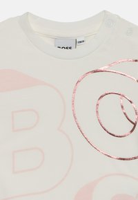 T-shirt de algodão branco com uma grande impressão em rosa claro da letra "B" e um design de espiral em dourado rosa brilhante. Fecho de botão de pressão no ombro.