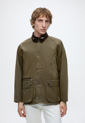 Jonge man draagt een olijfgroene jas met een zwarte kraag en grote voorzakken, gecombineerd met een witte broek, staand tegen een witte achtergrond.
