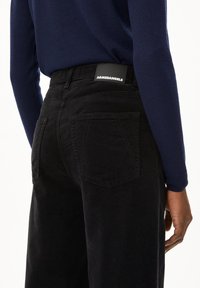Schwarze Cordhosen mit hoher Taille, auffälligen Gesäßtaschen und einem hinteren Etikett mit der Aufschrift "ARMEDANGELS." Die Textur ist gerippt und weich.