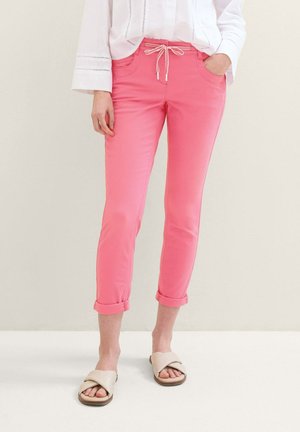 Femme portant un pantalon rose retroussé avec taille à cordon, un chemisier blanc à manches longues et des sandales beiges à lanières croisées, debout devant un fond uni.