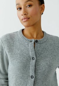 Vrouw in een zachte grijze gebuttonde cardigan met gouden ringoorbellen, die recht vooruit kijkt tegen een effen witte achtergrond.