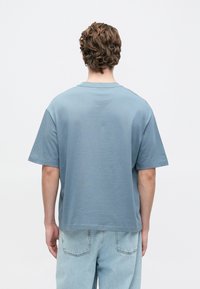 Vīrietis ar cirtainiem brūniem matiem, ģērbies brīvā zila t-kreklā un gaiši zilos džinos, stāv pretī vienkāršai baltai fona.