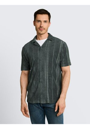 Homme portant une chemise à manches courtes gris foncé à boutons avec des rayures verticales et un jean bleu, se tenant devant un fond clair uni.