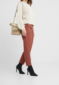 Frau in cremefarbenem Strickpullover, rostfarbenen Slim-Pants, schwarzen hochhackigen Stiefeletten und mit einer beigen Handtasche über der Schulter.