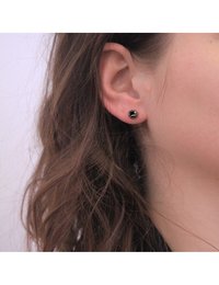 Orecchini a bottone rotondi con superficie in smalto nero, caratterizzati da un accento centrale in tono oro, posizionati su uno sfondo di capelli castani mossi.