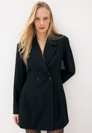 Femme aux longs cheveux blonds portant une robe blazer noire croisée, debout contre un mur blanc.