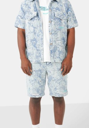 Man draagt een lichtblauw patroon kort mouw button-down shirt en bijpassende korte broek over een wit graphic T-shirt.