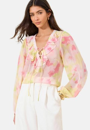 Frau mit langen dunklen Haaren, die eine transparente pastellfarbene Blumenbluse mit Rüschen und weiße, hoch taillierte Hosen trägt, accessoriert mit goldenen Armbändern und Ohrringen.