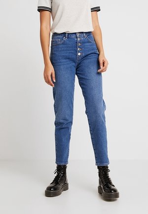 Jean bleu taille haute avec une fermeture à bouton, cinq poches et une coupe tapered. Associé à des bottes lacées noires et un haut gris.