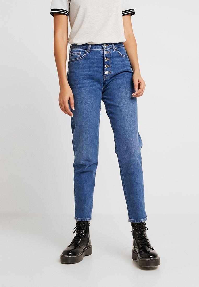 Jean bleu taille haute avec une fermeture à bouton, cinq poches et une coupe tapered. Associé à des bottes lacées noires et un haut gris.