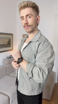 Ceas negru cu un cadran texturat, purtat la încheietura stângă, asortat cu o jachetă gri deschis. Fundalul include o canapea și lucrări de artă înrămate.