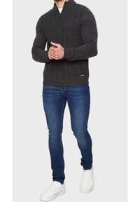 Maglione grigio scuro a maglia con colletto alto e chiusura con zip, abbinato a jeans blu slim-fit e sneakers bianche.
